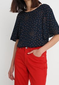 Marinebluse mit kleinen braunen Polka Dots, kurzen ausgestellt Ärmeln; kombiniert mit knallroten, eng anliegenden Jeans mit Fronttaschen und Gürtelschlaufen.