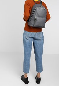 Sac à dos en cuir gris avec des fermetures éclair sur le dessus, matériel en argent et poche frontale. Porté avec un pull couleur rouille et un jean droit bleu clair.