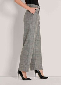 Frau in hoch taillierten grauen karierter weitem Bein Hose, mit Händen in den Taschen und schwarzen lackledernen Stöckelschuhen, die seitlich steht.