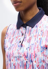 Γυναίκα που φοράει ένα αμάνικο, πολύχρωμο πουκάμισο πόλο με μοτίβο, με ναυτικό γιακά και κουμπιά, που φέρει το λογότυπο της Calvin Klein.