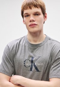 Jeune homme aux cheveux courts châtain clair portant un t-shirt gris Calvin Klein, bras croisés, expression neutre, fond clair uni.