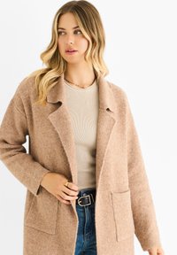 Beige wool blend long coat with a notched collar, open front, and two front pockets, worn over a fitted beige top and blue jeans.