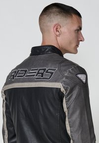 Muška motoristička jakna sa sivom teksturom i crnim kožnim detaljima, sa natpisom "RIDERS" izvezenim na leđima i logom na ramenu.