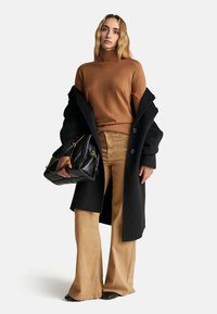 Schwarzer langer Mantel, brauner Rollkragenpullover, beigar farbene weite Hose, schwarze Handtasche mit goldenen Beschlägen. Glattes Material, taillierte Passform.