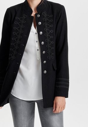 Sort jakke med indviklet broderi, sølvknapper og struktureret design. Kombineret med en hvid skjorte og grå jeans.