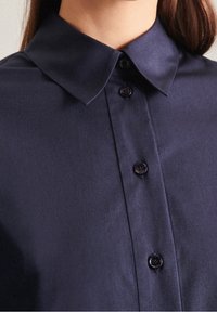 Námořnická modrá košile s límečkem vyrobená z hladkého materiálu, s knoflíkovou přední částí a tmavými knoflíky a strukturovaným výstřihem.