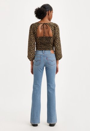 Frau mit lockigem Haar, die ein schwarzes langärmliges Oberteil mit Blumenmuster und offenem Rücken trägt, das am Hals gebunden ist, helle, hoch taillierte, gerade geschnittene Jeans und schwarze Schuhe.