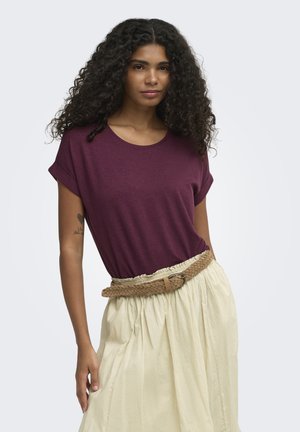 Mujer con cabello largo y rizado que lleva una camiseta de manga corta burdeos metida en una falda beige de talle alto con un cinturón trenzado.