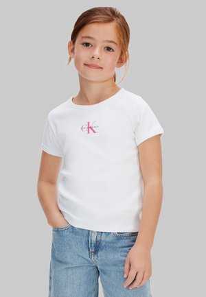 Jeune fille aux cheveux roux portant un t-shirt blanc Calvin Klein et un jean bleu, debout avec une main dans la poche devant un fond gris.