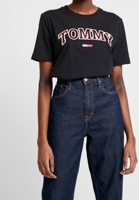 Camiseta de algodón negra con "TOMMY" bordado en rojo, blanco y verde. Combinada con jeans de tiro alto en azul oscuro con textura de denim clásica.