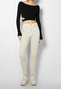 Mujer con una camiseta negra de manga larga corta con corte y unos pantalones ajustados beige de cintura alta, combinados con zapatillas de plataforma.