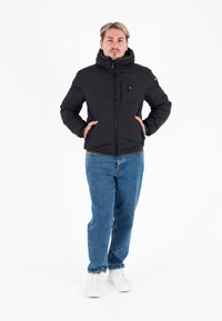 Giacca nera imbottita con colletto alto, tasche frontali e dettaglio del logo giallo, indossata con jeans blu e sneakers bianche.