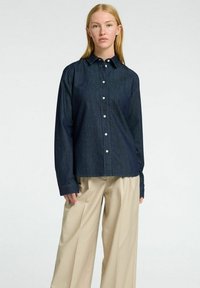 Tamsiai mėlynas džinsinis marškinis su klasikinėmis apykaklėmis, ilgomis rankovėmis ir baltais mygtukais, derinamas su šviesiai smėliniais plačiakulkšniais kelnais.