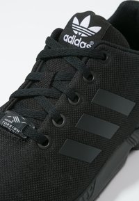 Black athletic shoes with textured mesh upper, black tonal three stripes, and textured laces. Features a branded black tag and rubber sole.