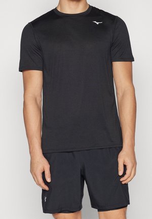 Mies, joka pitää mustaa lyhythihaista urheilupaitaa ja mustia shortseja, seisoo rentoina käsivarret sivuilla yksiväristä taustaa vasten.