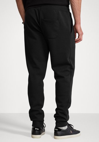 Black sweatpants with a straight cut, elastic waistband, and a rear pocket. Paired with dark sneakers featuring a logo on the heel.