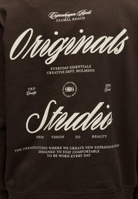 Sweat-shirt marron avec texte blanc indiquant "Originals Studio" et petites phrases sur la créativité, le confort et les détails du port quotidien.