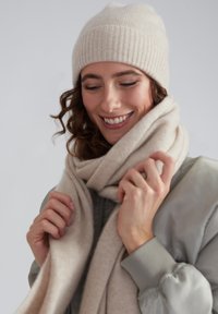 Beige gerippte Strickmütze und weicher beiger Schal, der um den Hals gewickelt ist. Der Schal hat eine glatte Textur und die Mütze sitzt eng.