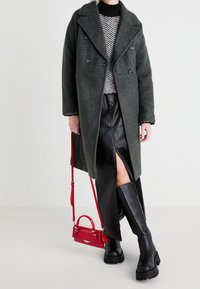 Abrigo verde de doble botonadura, tejido texturizado, suéter de patrón en blanco y negro, falda de cuero negra con abertura, botas negras gruesas, bolso pequeño rojo.