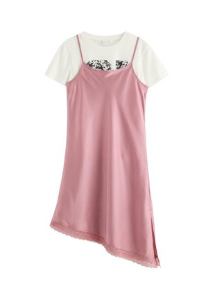 Ung pige med langt brunt hår iført en pink kjole over en hvid skjorte, beige cardigan, hvide sokker og hvide sneakers, stående på en ensfarvet baggrund.