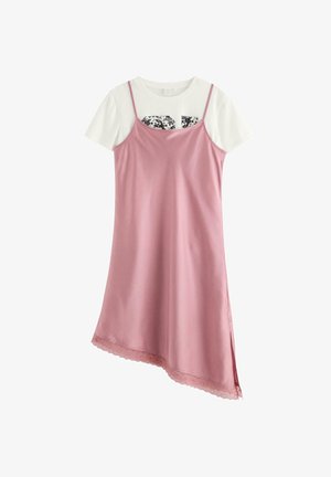 Ung pige med langt brunt hår iført en pink kjole over en hvid skjorte, beige cardigan, hvide sokker og hvide sneakers, stående på en ensfarvet baggrund.