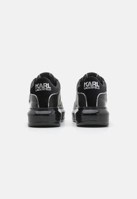 Black sneakers with a glossy finish, featuring a padded collar, white stitching, and a logo on the heel. Low-top design with a thick sole.