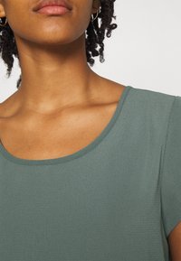 Zielona, teksturowana bluzka z okrągłym dekoltem, z krótkimi rękawami i subtelnymi detalami w szwie. Gładki materiał z lekkim połyskiem.