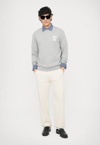 Szara bluza z okrągłym dekoltem i małym logo, noszona na niebieskiej koszuli z kołnierzykiem. Białe spodnie i czarne loafersy dopełniają strój.