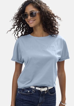 Frau mit lockigem Haar, die Sonnenbrille trägt, ein hellblaues "BONJOUR"-T-Shirt, dunkle Jeans mit weißem Gürtel und ein silbernes Armband trägt, leicht lächelnd.