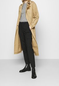 Beige lange jas over grijze t-shirt, gecombineerd met zwarte cargobroek en zwarte enkellaarsjes. Soepele stoffen en knoopaccenten zichtbaar.