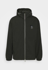 Svart vindjacka med dragkedja, justerbar huva, elastiska ärmmuddar och en märkeslogga på vänster framsida.