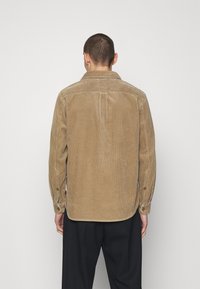 Camisa de pana color camel con cuello puntiagudo y cierre de botones, presenta costillas verticales y un dobladillo ligeramente curvado. Tejido transpirable y texturizado.