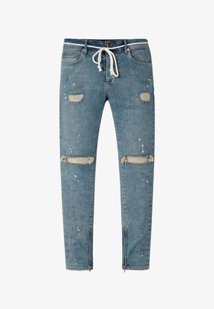 Jean slim bleu en denim avec taille à cordon, patchs usés sur les cuisses et les genoux, éclaboussures de peinture blanche et fermetures éclair aux chevilles.