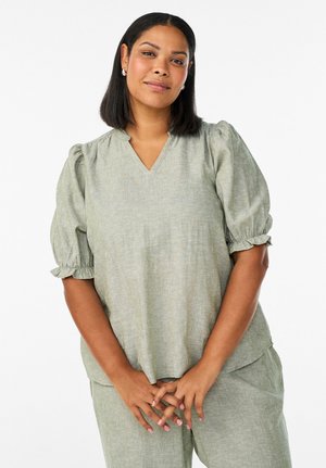 Ženska s črnimi srednje dolgimi lasmi, oblečena v svetlo zeleno bluzo z napihnjenimi rokavi in ujemajoče hlače, stoji z rokami zaprtimi spredaj.