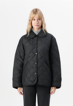 Femme portant une veste matelassée noire avec un grand col et des boutons-pression, associée à un pull à col roulé gris et un pantalon foncé.