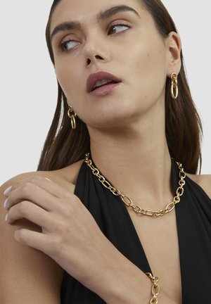 Woman wearing linked gold chain jewelry set including necklace, bracelet, and oval hoop earrings, with black sleeveless top and light makeup.