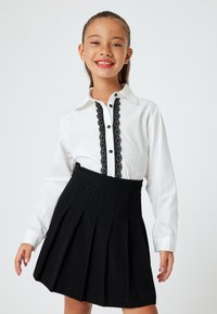 Camicia bianca con bottoni neri e bordo in pizzo, abbinata a una gonna nera plissettata con vita e texture liscia.