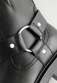 Black leather boot featuring a textured finish, silver circular hardware, and a wide strap with metal studs. Detailed stitching accents the design.