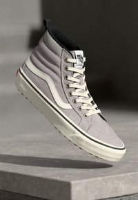 High-top sneaker in light gray suede with white accents, cream laces, and signature logo label on the tongue; textured side panels.