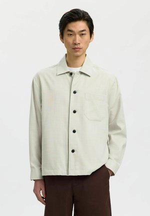 Joven con una chaqueta de camisa color beige claro con botones negros y un bolsillo en el pecho, combinado con pantalones marrón oscuro, de pie sobre un fondo blanco.