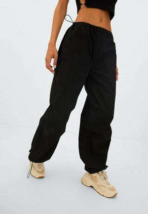 Vrouw draagt zwarte wijde broekspijpen, beige sneakers en een zwart cropped topje met een hartvormige ring op haar navel.