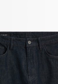 Dark denim jeans with a classic five-pocket design, featuring a metal button closure and slight texture. Label visible at the waistband.