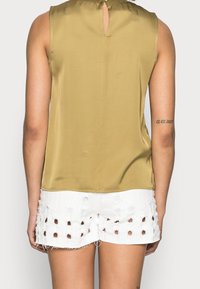 Top sin mangas verde oliva con un corte en forma de llave en la espalda. Combinado con pantalones cortos blancos desgastados con patrones recortados y bordes deshilachados.