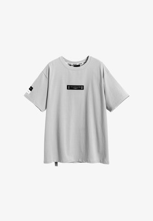 Camiseta de algodón gris claro, corte relajado, cuello redondo, mangas cortas, con una etiqueta rectangular negra en el frente y acentos laterales.