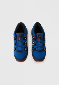 Μπλε αθλητικά παπούτσια με υφασμένο επάνω μέρος, μαύρες και λευκές λεπτομέρειες, ελαστικά κορδόνια και κόκκινο κουμπί ρύθμισης. Εξωτερική σόλα από καουτσούκ με αντιολισθητικό σχέδιο.