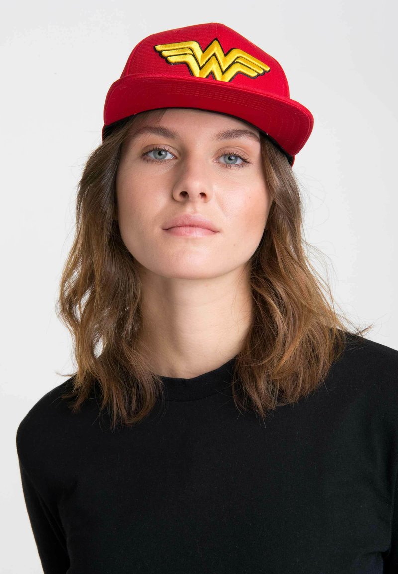 Red baseball cap with yellow Wonder Woman logo on the front, featuring a flat brim and structured design. Worn with a black long-sleeve top.