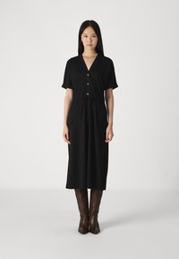 Vestido midi preto com mangas curtas, decote em V, detalhe de botões, cinto atado na cintura e dois bolsos frontais. Usado com botas castanhas até ao joelho.