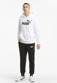 Hvid hætte sweatshirt med sort "PUMA" logo, parret med sorte bukser med et lille hvidt logo. Modellen bærer hvide sneakers.