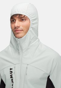 Ljusgrå huvjacka med hög krage, som har två fickor med dragkedja och svarta detaljer; tillverkad av ett slätt, elastiskt material.
