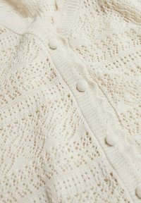 Gilet en tricot crème avec motif en dentelle en losanges et boutons recouverts de tissu sur toute la longueur de la patte de boutonnage.